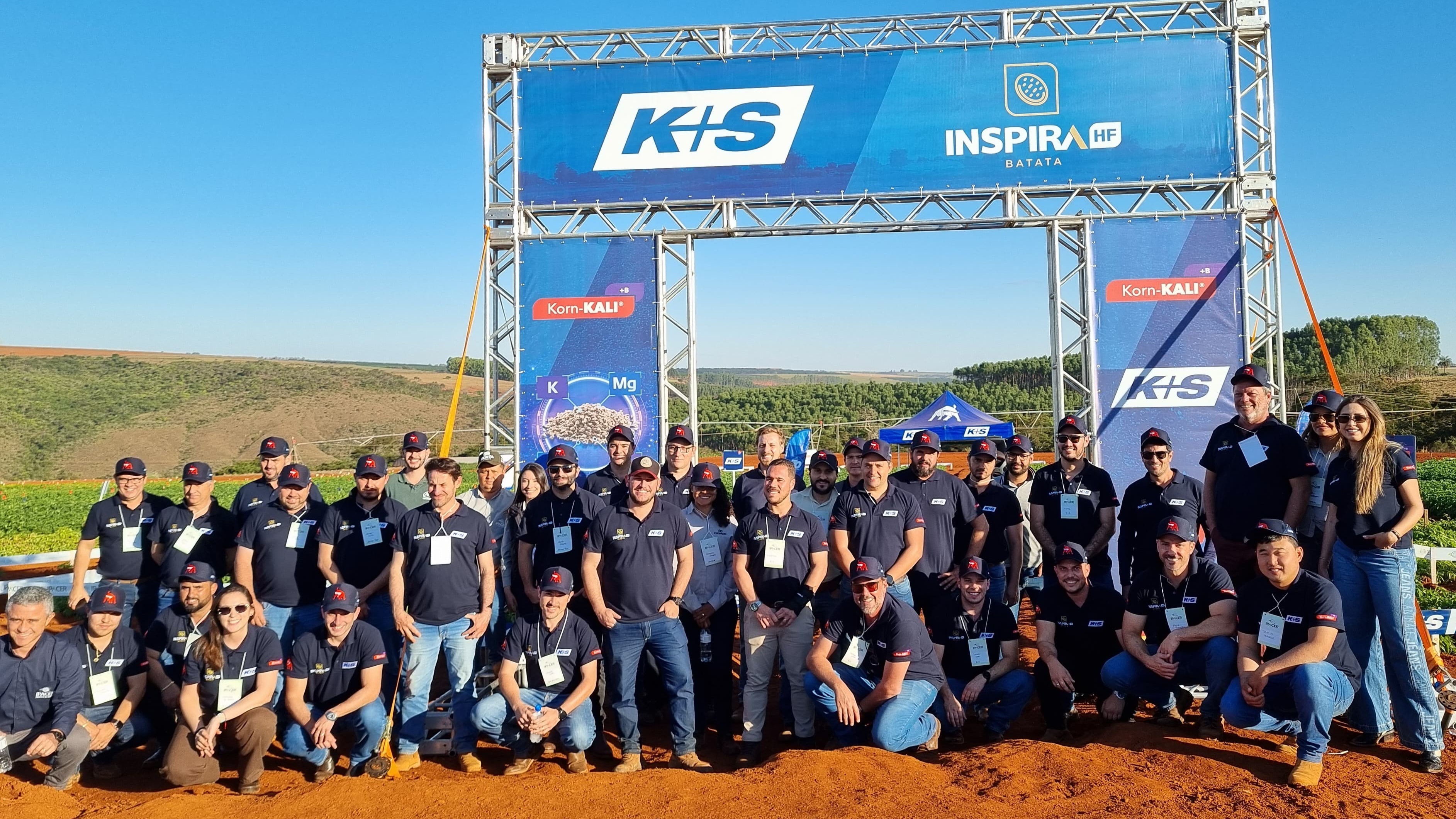 Grupo de pessoas posando para foto em frente a uma estrutura metálica com banners da K+S e do projeto Inspira HF Batata. Ao fundo, há uma área agrícola com vegetação e céu parcialmente nublado. No canto superior direito, aparece o logotipo da K+S.